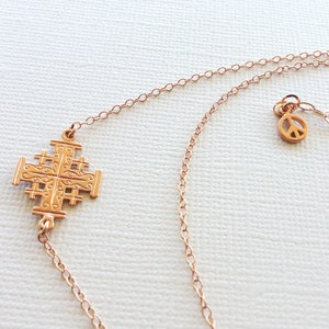 May include: A gold necklace with a Jerusalem cross pendant and a peace sign charm. The necklace is made of a delicate chain.