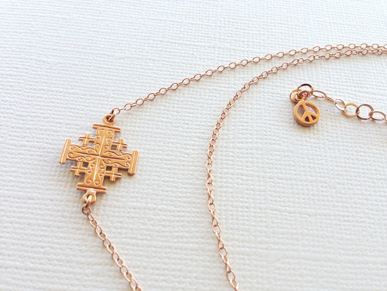 May include: A gold necklace with a Jerusalem cross pendant and a peace sign charm. The necklace is made of a delicate chain.
