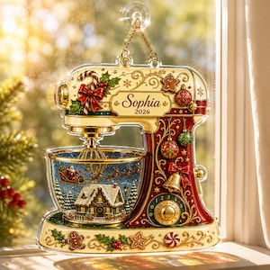 May include: A decorative acrylic stand mixer ornament featuring a cream and red color scheme with gold accents. The mixer displays the name Sophia and the year 2026. The mixing bowl contains a winter scene with a house, trees, and Santa in a sleigh.