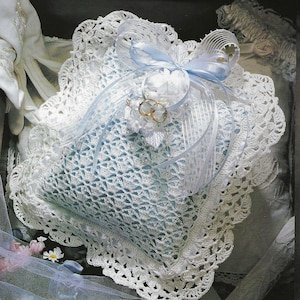 May include: A white crocheted ring bearer pillow with a blue satin ribbon and two gold rings. The pillow is decorated with a white lace trim.