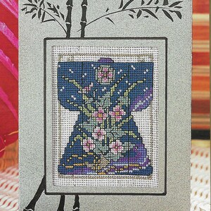 May include: Cross stitch pattern featuring a blue and purple kimono with pink flowers and green leaves. The pattern is framed with a black bamboo design and the text "Cross Stitch Greetings" and "Cartes de Souhait en Point de Croix".
