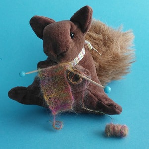 May include: A brown plush squirrel with a long, fluffy tail is holding knitting needles and a colorful knitted scarf. The squirrel is wearing a striped ribbon around its neck.