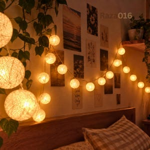 Pode incluir: Um fio de luzes esféricas de algodão tecido que emitem um brilho quente e suave. As luzes estão dispostas em cascata sobre uma parede decorada com várias impressões retangulares e o texto Good Vibes Only, apresentando um ícone de coração.