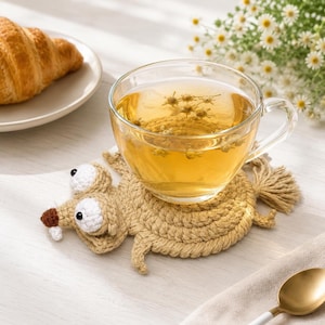 May include: A clear glass teacup filled with golden herbal tea containing small flowers, resting on a tan crocheted coaster shaped like a squirrel with large white eyes, a brown nose, and a bushy tail, positioned next to a croissant.