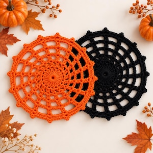 Peut inclure: Deux napperons circulaires au crochet avec un motif de toile d'araignée, l'un orange vif et l'autre noir. Les napperons sont disposés avec des citrouilles orange et des feuilles d'automne séchées éparpillées sur une surface blanche.