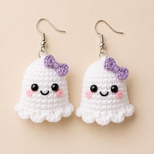 Peut inclure: Une paire de boucles d'oreilles fantômes en crochet blanc avec des yeux en perles noires, un sourire noir brodé, des touches de blush rose et un petit nœud en crochet violet sur le côté. Les boucles pendent à des crochets en métal argenté.