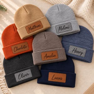 May include: A collection of seven knit beanies in various colors including tan, gray, orange, dark gray, brown, blue, and navy. Each beanie features a rectangular leather or felt patch with a name engraved in black script: Matthias, Meloni, Charlotte, Amelia, Henry, Oliver, and Lucas.