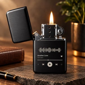 May include: A matte black metal flip-top lighter with a lit flame. The front features a white graphic of a music player interface, including a sound wave, the text Another Love by Tom Odell, a heart icon, and playback control buttons.