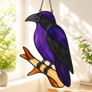 May include: A stained glass suncatcher shaped like a crow perched on a branch. The bird features deep purple and black glass panels with a dark beak. The branch is constructed from amber and tan textured glass pieces, all held together by black metal.