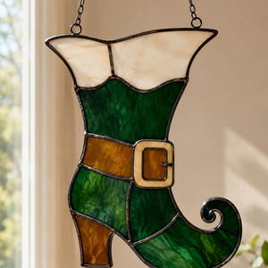 Pode incluir: Um apanhador de sol de vitral em forma de bota com biqueira enrolada. A bota apresenta um punho branco, um corpo verde e um salto e correia de cinto castanhos com uma fivela clara. Toda a peça é unida por linhas de soldadura de metal escuro.
