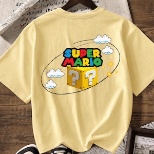 May include: A pale yellow short-sleeve t-shirt featuring a graphic print of the Super Mario logo above a yellow question mark block. The design includes pixelated white clouds and yellow star icons enclosed within a thin black oval outline.