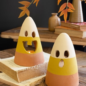 May include: Two cone-shaped ceramic candle holders painted in horizontal stripes of white, yellow, and orange to resemble candy corn. Each holder features two oval eye cutouts and a central mouth opening, with one containing a lit tea light candle.