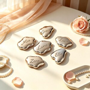 May include: Six irregular, cloud-shaped gold-toned compact mirrors, each engraved with a unique name: Grace, Mia, Luna, Luna, Ella, and Zara. The metallic surfaces are polished and reflective, arranged on a smooth surface with scattered pearls.