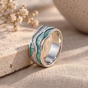 May include: A silver-toned ring featuring a wavy, multi-layered design with inlaid turquoise-colored enamel and a textured, shimmering silver-toned surface. The band has a polished metallic finish and a smooth, rounded interior profile.
