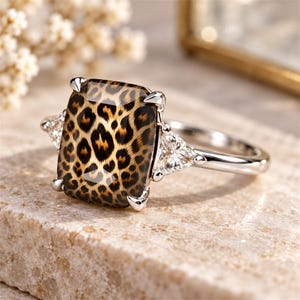 May include: A silver-toned ring featuring a large, rectangular, faceted centre stone with a brown and black leopard print pattern. The centre stone is held by four prongs and is flanked by two smaller, clear, triangular-cut side stones.