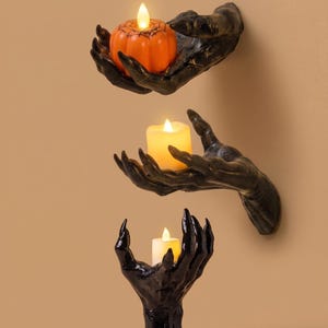 May include: Three black wall-mounted or standing hand sculptures with long, pointed fingers. Two hands are mounted horizontally, one holding an orange pumpkin-shaped LED candle with a spiderweb design, and one holding a plain white LED candle.
