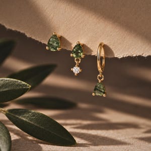 May include: Three gold earrings featuring green pear-shaped stones. One is a stud, one is a stud with a small clear round stone drop, and one is a small hoop with a green pear-shaped stone drop. The metal is yellow gold with a polished finish.