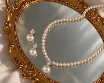 Op de afbeelding: Een sieradenset bestaande uit een parelketting en twee bijpassende oorbellen. De ketting heeft een enkele rij witte parels met een druppelvormige hanger met een heldere steen. Elke oorbel is voorzien van een bijpassende parel en steen.