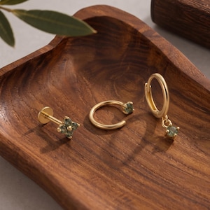 Könnte beinhalten: Ein Set aus drei goldfarbenen Schmuckstücken mit kleinen, facettierten olivgrünen Steinen. Die Kollektion umfasst einen Stecker mit flacher Rückseite und vierblütigem Steinmotiv, einen einfachen offenen Ring und eine kleine Klappcreole mit einem Anhänger.