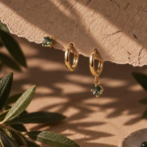 Puede incluir: Un conjunto de joyas de oro que consta de un pendiente de botón con una piedra preciosa verde redonda, un pendiente de aro de oro liso y un pendiente de aro de oro con una piedra preciosa verde redonda colgante, sobre una superficie beige texturizada.