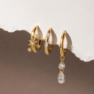 May include: Three gold hoop earrings. The first features a starburst design with small clear stones. The second is a plain hoop set with rectangular baguette-cut clear stones. The third is a plain hoop with a dangling round and pear-shaped clear stone.