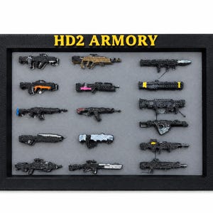 Może przedstawiać: Czarna prostokątna ramka ekspozycyjna z żółtym napisem HD2 ARMORY u góry. Wewnątrz znajduje się piętnaście miniaturowych modeli broni ułożonych w siatkę na szarym tle, charakteryzujących się różnymi kształtami, kolorami i teksturami.