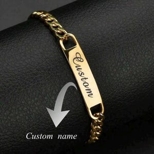Peut inclure: Un bracelet à chaîne en métal doré doté d'une plaque d'identification rectangulaire centrale. La plaque est gravée du mot Custom en écriture cursive. Une flèche blanche pointe vers la plaque, accompagnée du texte Custom name.