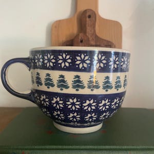 May include: A ceramic mug featuring a dark blue and cream color palette with a repeating pattern of white snowflakes on the top and bottom bands and a central row of green pine trees. The mug has a curved handle and a wide, rounded bowl shape.