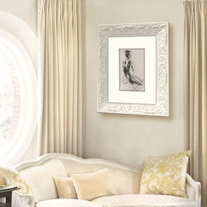 May include: A black and white drawing of a nude figure in a white frame with an ornate design. The drawing is hanging on a white wall in a room with white curtains and a white couch.