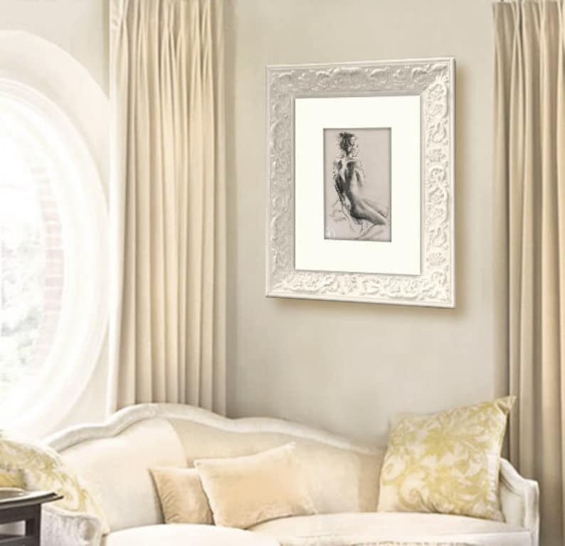 May include: A black and white drawing of a nude figure in a white frame with an ornate design. The drawing is hanging on a white wall in a room with white curtains and a white couch.