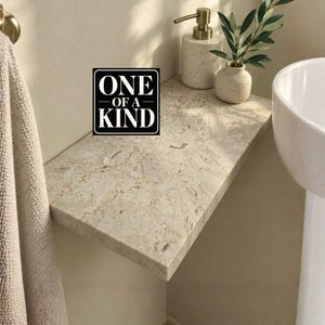 Pode incluir: Uma prateleira retangular de pedra bege com um padrão natural salpicado. Na prateleira, há um dispensador de sabonete cor de creme com uma bomba dourada e um pequeno vaso contendo uma planta verde. Uma placa preta com texto branco diz ONE OF A KIND.
