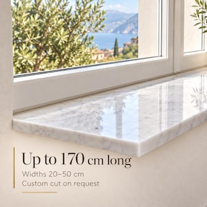 May include: A polished white marble window sill with grey veining. The text on the image states: Up to 170 cm long, Widths 20-50 cm, Custom cut on request. The sill is installed beneath a window with a view of a coastal landscape.