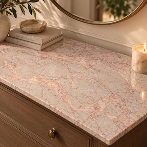 Peut inclure: Une commode en bois à deux tiroirs avec des boutons ronds en laiton et un plateau en marbre rose poli. Sur le dessus sont posés deux livres empilés intitulés The Kinfolk Home et At Home, un petit bol en pierre, un vase blanc et une bougie.