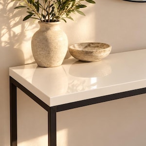Peut inclure: Une console rectangulaire dotée d'une surface supérieure blanche lisse et brillante, soutenue par un cadre fin en métal noir. La table est positionnée horizontalement, présentant un design linéaire épuré avec quatre pieds et une base structurelle.