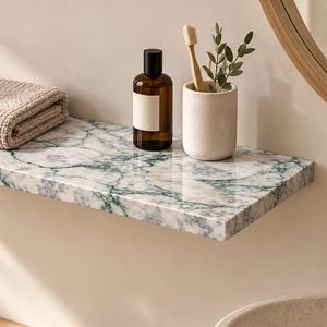 Op de afbeelding: Een rechthoekige wandplank met een wit en groen marmerpatroon. Op de plank ligt een gevouwen beige handdoek met wafelstructuur, een donker amberkleurige glazen fles met een leeg wit etiket en een matte witte beker met een houten tandenborstel.