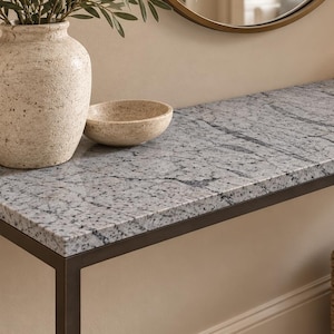 Pode incluir: Uma mesa consola retangular com um tampo de granito salpicado de cinzento e branco, suportada por uma estrutura metálica fina e escura. A superfície da mesa contém um vaso bege texturado com vegetação, uma pequena taça bege e livros.