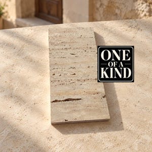 May include: A rectangular piece of beige travertine stone with a porous, textured surface and natural linear veining. A black square graphic with white text reads ONE OF A KIND, positioned to the right of the stone tile on a matching surface.