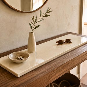 Op de afbeelding: Een rechthoekig, crèmekleurig stenen dienblad staat op een console-tafel van donker hout. Op het dienblad staan een kleine beige keramische kom met metalen sleutels, een slanke beige vaas met een groen takje en een zonnebril.