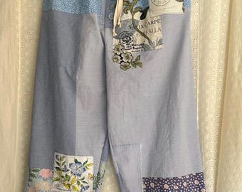 Op de afbeelding: Blauw-wit gestreepte patchworkbroek met bloemen- en patroonstoffen patches. De broek heeft een taille met trekkoord en hangt aan een hanger. Patches zijn onder meer bloemen- en vintage postzegelontwerpen.