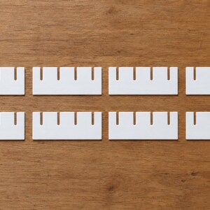 May include: Eight rectangular white plastic display cards arranged in two rows. Each card features four evenly spaced vertical notches along the top edge, designed for holding items securely in place for organized presentation.