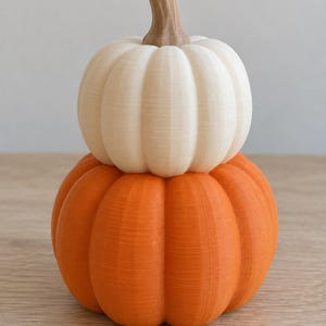 May include: A stacked decorative pumpkin ornament featuring a large orange pumpkin base topped with a smaller cream-colored pumpkin. Both pumpkins have vertical ridges and a brown stem at the top, showcasing a layered, textured finish.