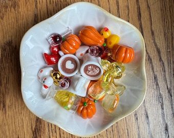 May include: A white, scalloped-edge bowl filled with miniature resin charms including orange pumpkins, white coffee mugs with latte art, yellow rubber ducks, clear glasses with liquid designs, and small red and white bird figures.