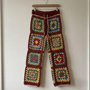 May include: Crochet pants featuring a patchwork design of multi-colored granny squares in shades of red, yellow, blue, green, and cream. The trousers have a dark red ribbed waistband with a matching drawstring and a straight-leg silhouette.