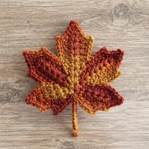 Op de afbeelding: Een gehaakte esdoornblad-applicatie met een verloop van warme herfstkleuren, variërend van diep bordeaux en donkerrood aan de buitenranden tot een levendig goudgeel en oranje naar het midden toe, compleet met een klein steeltje aan de onderkant.