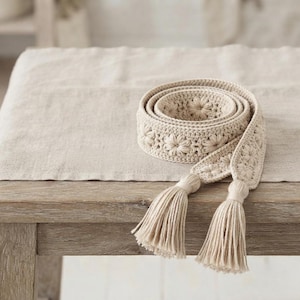 May include: A beige crochet belt featuring a repeating floral pattern throughout the length of the band. The belt is coiled and finished with two long, matching beige tassels at the ends, constructed from a series of interconnected yarn loops.