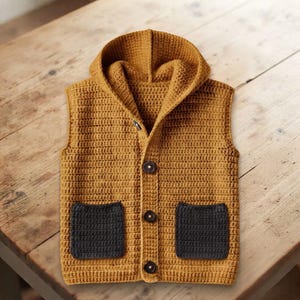 May include: A mustard yellow sleeveless hooded vest made of crocheted yarn. The garment features a front button closure with three dark brown buttons and two contrasting dark gray square pockets positioned on the lower front of the vest.