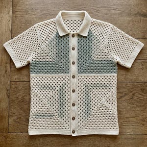 May include: A short-sleeved polo shirt crafted from cream and sage green crochet fabric. The garment features a classic collar, a front button placket with eight brown buttons, and a repeating square-based openwork pattern throughout the design.
