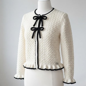 May include: A cream-colored long-sleeve cardigan featuring a textured knit pattern, a round neckline, and a front closure with two black bows. The garment includes black trim along the edges, cuffs, and a ruffled hem, with a small label at the waist.