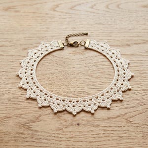 May include: A cream-colored crochet lace choker necklace featuring a scalloped edge pattern. The necklace is secured with a bronze-toned metal lobster claw clasp and a short extender chain, providing a secure and adjustable fit for the wearer.