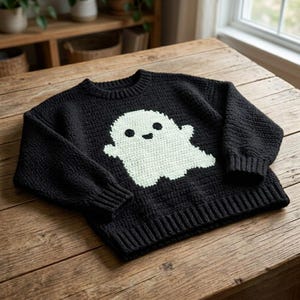 May include: A black long-sleeve sweater featuring a large, centered, pale green ghost graphic. The sweater is constructed with a textured, knit-like pattern, including a ribbed crew neckline, ribbed cuffs, and a ribbed hem at the bottom edge.